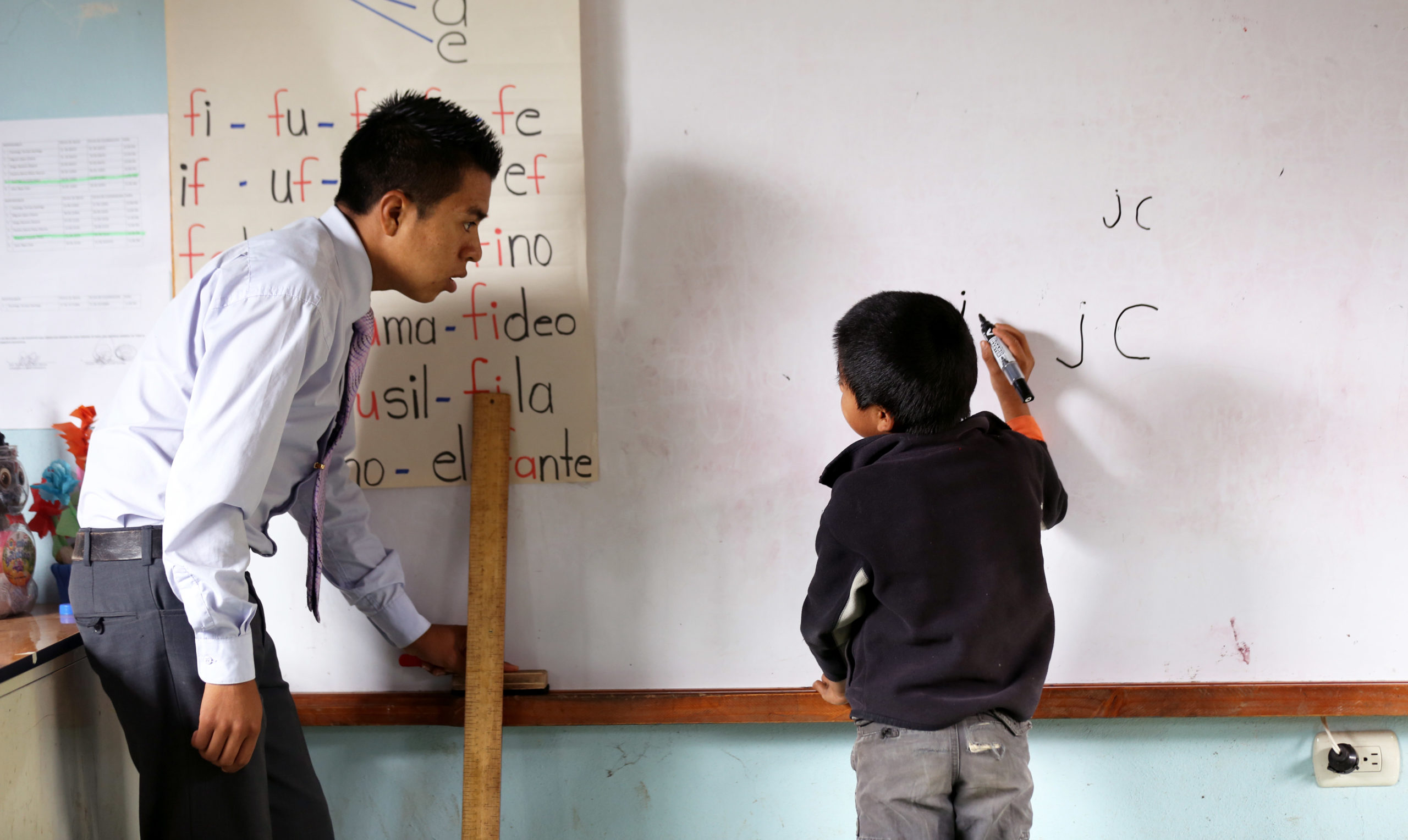 A teacher helps a student write on the white board at the front of a classroom in Guatemala.