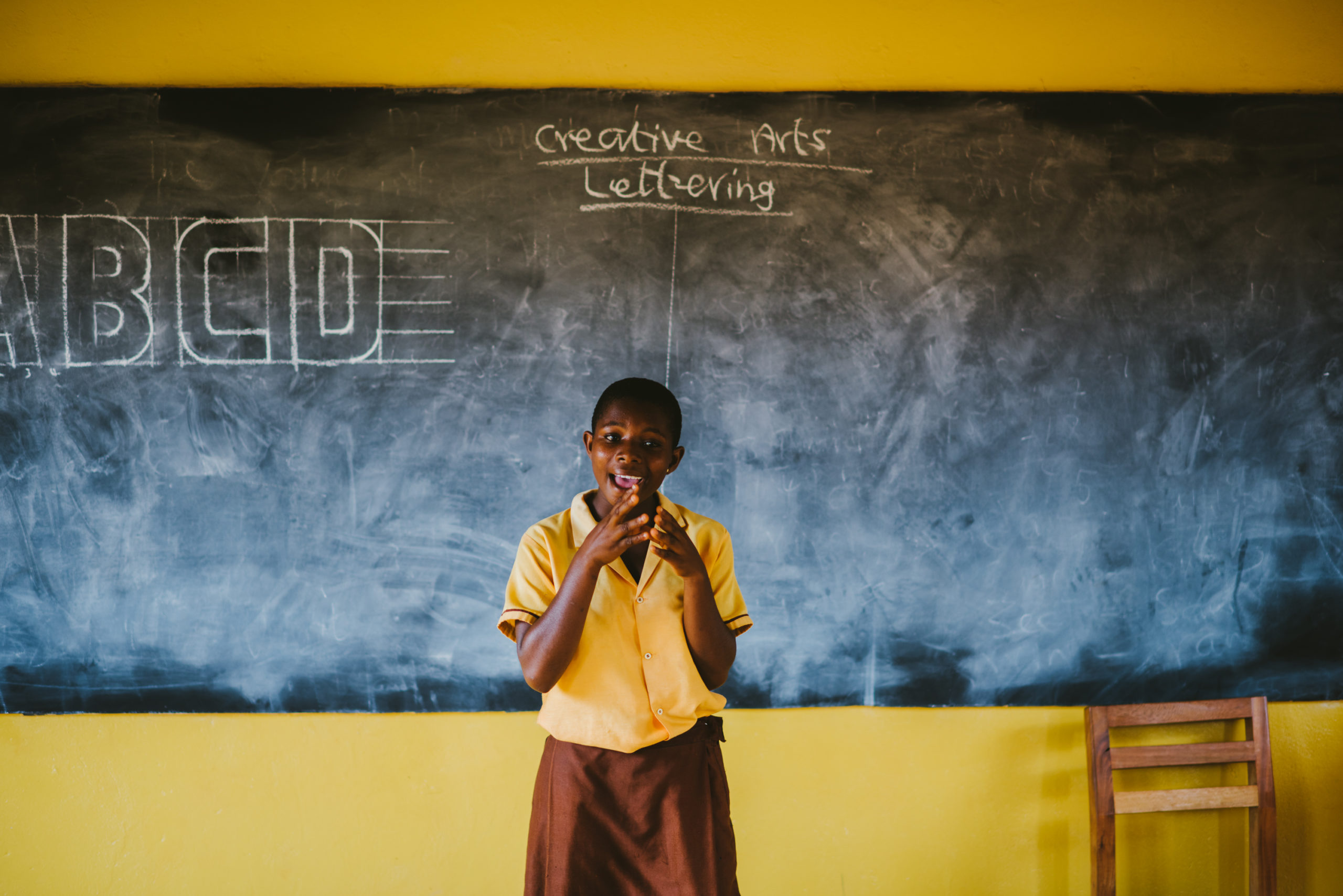 A student in Ghana stands in front of the blackboard inside a PoP-built classroom.