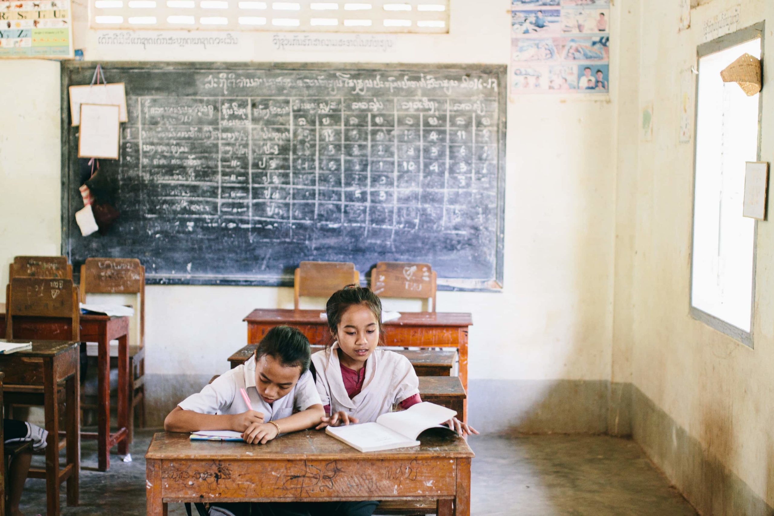 Two students share a desk inside a classroom in Laos with a chalkboard on the wall behind them.